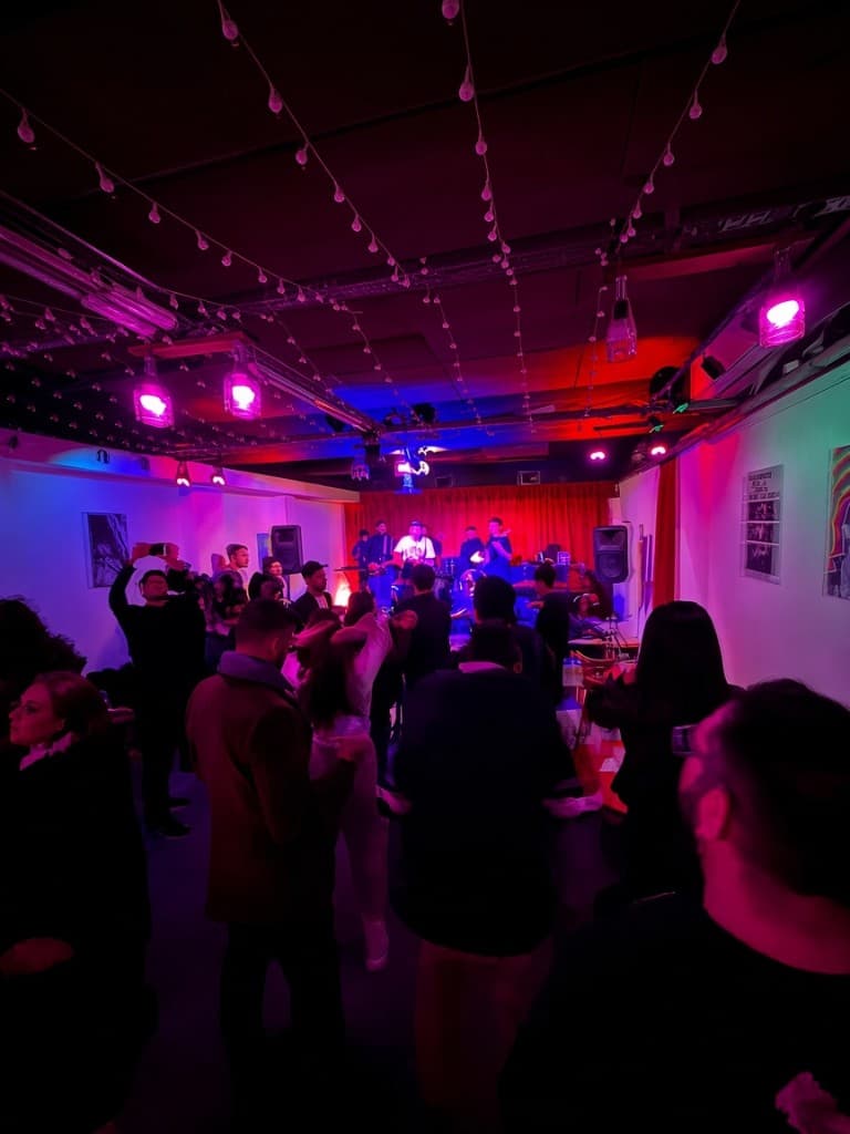 Sala llena de La Llama en un show: banda en vivo sobre el escenario con cortina roja, público de pie y guirnaldas de luces cruzando el techo.